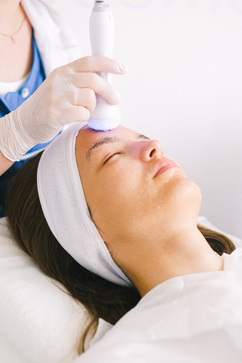 Professional female beautician in gloves and robe using ultrasound apparatus to clean female customer face lying with eyes closed in spa center